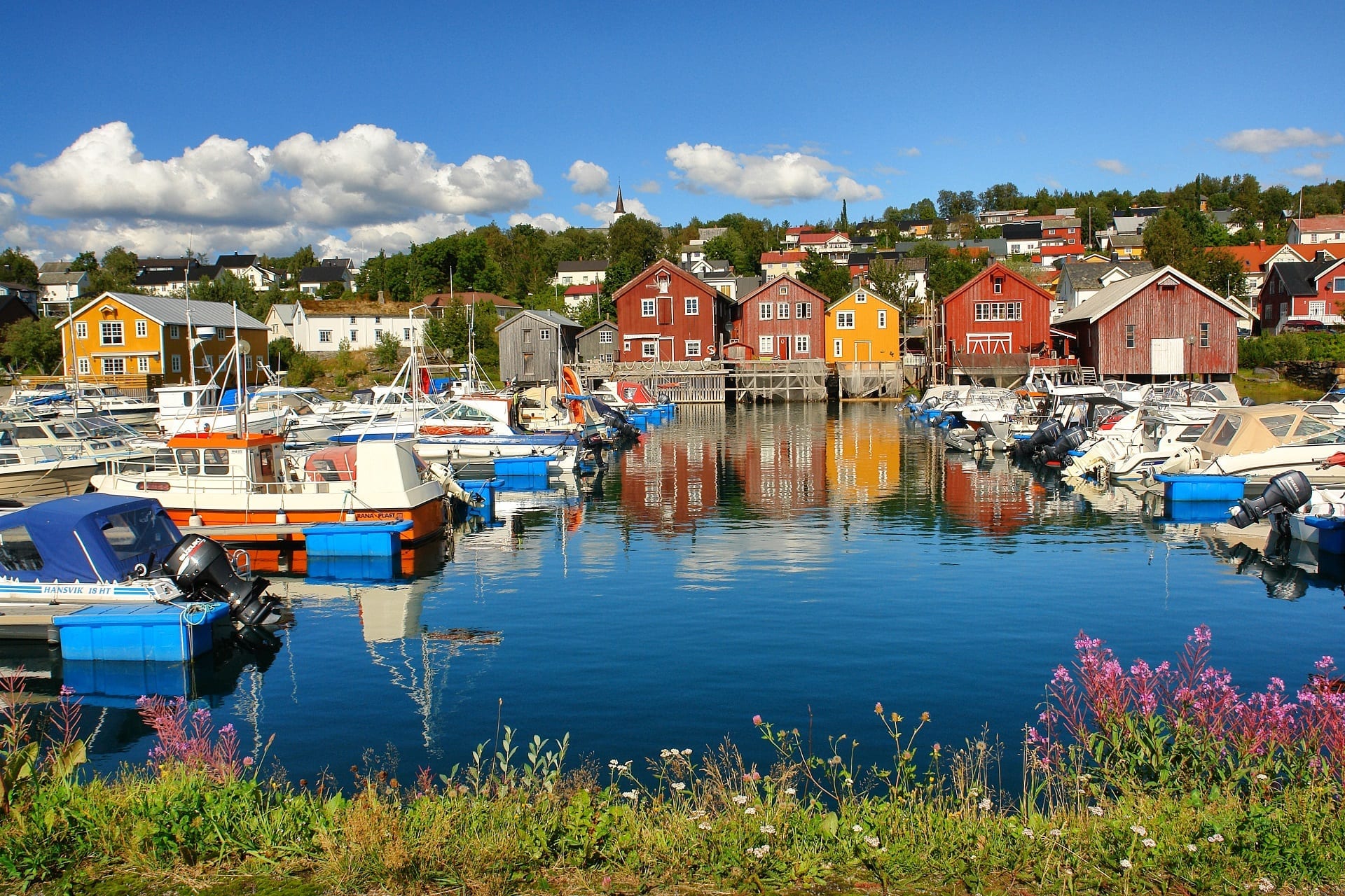 Beautiful and colorful photo of boat houses next to the marina at Hemnesberget with boats and flowers in the foreground.