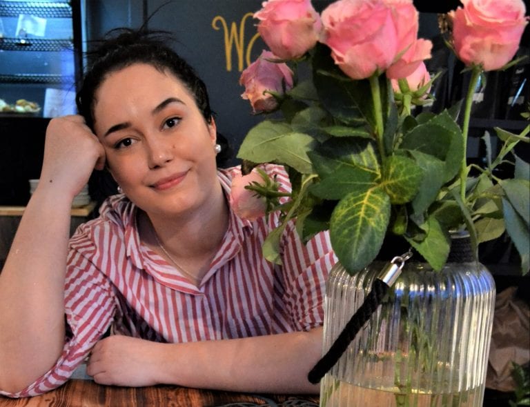 Smiling and colorful Sandra, standing behind the counter decorated with roses at her coffee shop, “Lille Havfruen Kafe”.