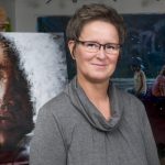 Artist, Yvonne, smiling with her glasses on and standing in front some of her beautiful paintings.