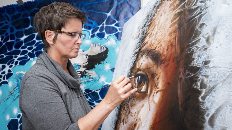 Artist, Yvonne, concentrated while painting the eye of a girl on one of her paintings. Another painting is seen in the back.