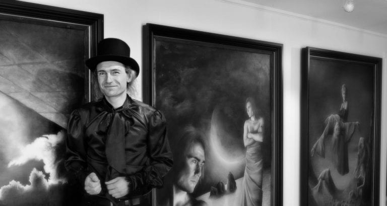 Black-and white photo of the smiling artist, Sigmund, in front of some of his paintings at an exhibition.