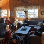 People sitting in a cozy living-room inside a cabin in the mountains with the sun shining in through the windows.