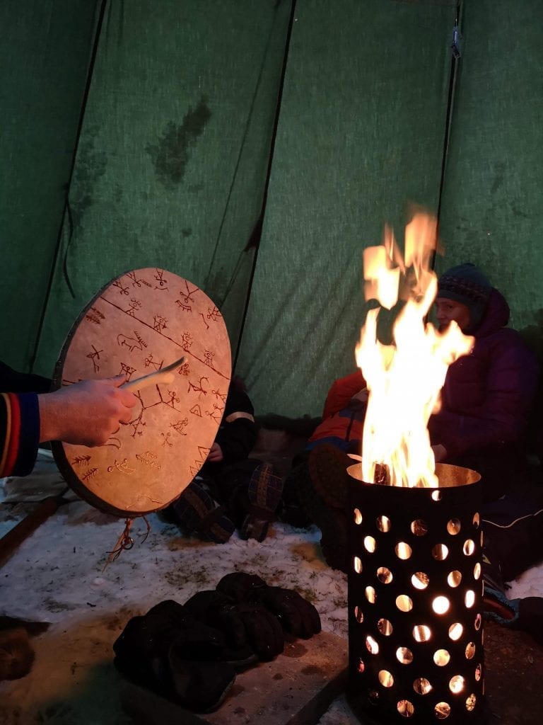 Someone playing on a sami drum covered with sami pictograms in front of a fire inside a lavvo, a sami tent.