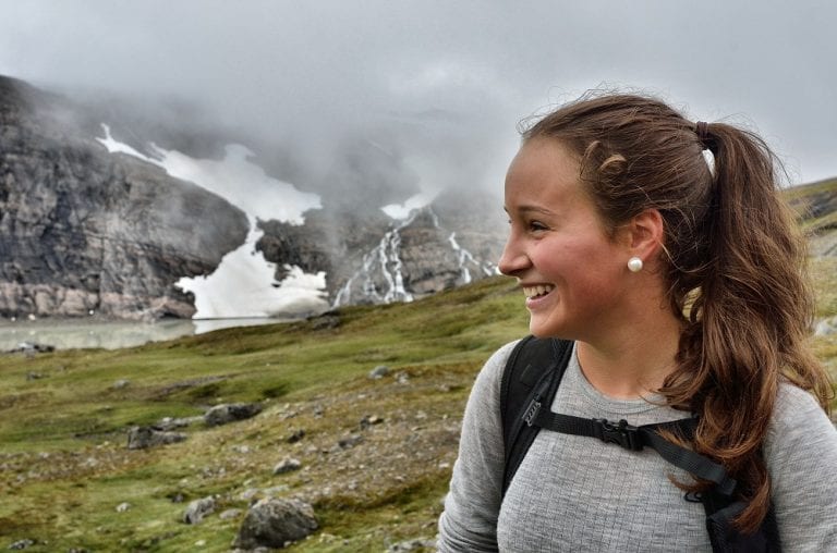 Happy young girl hiking in Okstindan on a cloudy day with snow and a small mountain lake seen in the background.