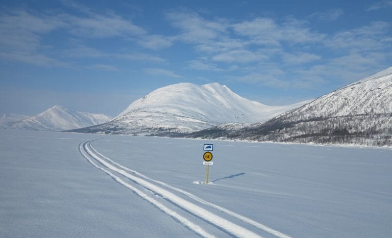 Snowmobile track with a speed sign next to it, in the middle of an open, white landscape surrounded by majestic mountains.