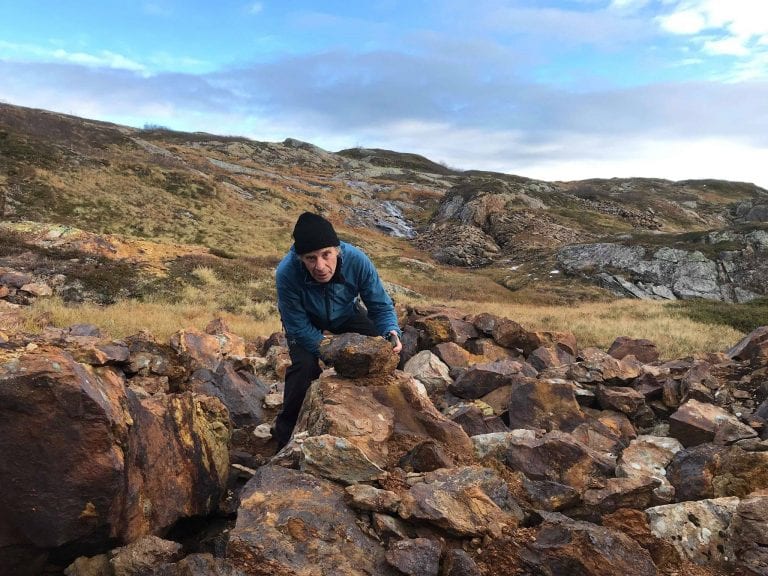 Man standing between rocks in a mountain landscape while having his hands on one rock in particular.