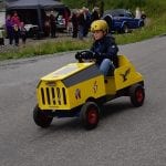 Young boy with a yellow helmet driving a small home-made yellow car down the road with people watching him.