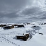 Six wooden cabins surrounded by snow in a mountain landscape.