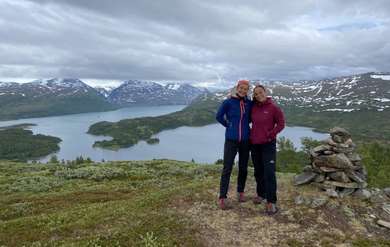 Two women hiking in front of a lake and tall mountains in the horizon