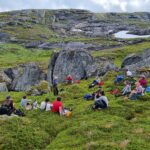 About 15 people sitting on the ground in a green environement necxt to the big boulders named after the sami Stor Nila