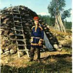 A sami with a red hat in front of a traditional hut