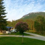 Small cabins surrounded by green and lively nature right next to the river Røssåga and the mountain Klubben in the back.