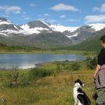 Hiker with her dog next to the lake at Stekvasselv Farm with a great view over snow-covered mountain tops in the horizon.