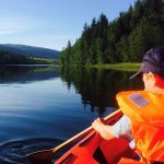 Young boy in a bright orange life jacket paddling in a canoe on Røssåga river, on a quiet and sunny day.