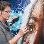 Artist, Yvonne, concentrated while painting the eye of a girl on one of her paintings. Another painting is seen in the back.