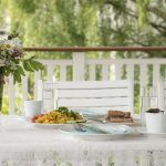 Colorful brunch outside on a white table decorated with flowers, on a white porch in green surroundings.