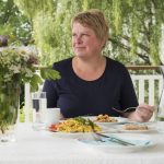 Woman eating a colorful brunch outside on a white table decorated with flowers, on a white porch in green surroundings.