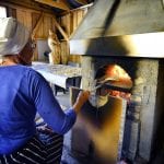 Elderly lady in blue sitting in front of baking oven with “kamkaker”, local bread, ready to be baked.