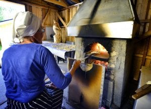 Elderly lady in blue sitting in front of baking oven with “kamkaker”, local bread, ready to be baked.