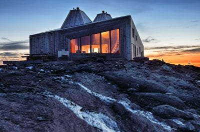 Rabothytta seen from the outside, lit up by orange lights inside that matches with the orange sunset outside.