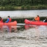 Four young people in two red canoes with life vests on, on the clear-watered river Røssåga on a nice summer day.