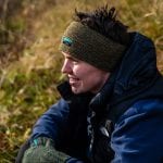 Smiling man sitting outside in nature wearing a matching set of green knitted headband and mittens from “Okstind”.