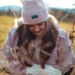 Smiling woman outside in nature wearing a purple winter hat matching her jacket, and white mittens, both from “Okstind”.