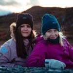 Two smiling women in colorful jackets outside in nature, with winter hats in brown and blue, and mittens from “Okstind”.