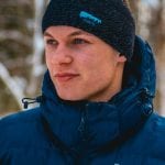 Smiling man outside in nature wearing a dark blue knitted headband from “Okstind”, that matches with his blue jacket.