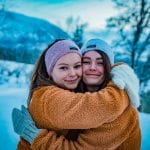 Two sisters smiling and hugging each other, wearing knitted headbands and mittens from “Okstind”.