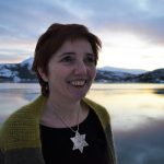 Portrait of goldsmith Merete wearing one of her necklaces in the shape of a star, standing in front of the beautiful fjord.