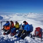Skiers sitting in colorful clothes, taking a break in snowy, white mountain surroundings on a clear blue-skied day.