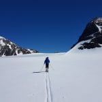 Skiers in an open and white mountain landscape walking towards big mountains on a blue-skied day.