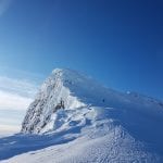 Skier climbing a steep and rocky mountain covered with snow on a blue-skied day.