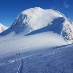 Skiers climbing a white mountain surrounded by other big, majestic mountains on a blue-skied day.