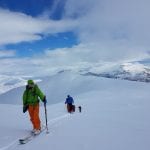 Skiers in colorful clothes climbing a white and open mountain landscape, surrounded by other mountains.