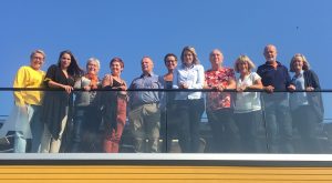 11 smiling partners of the Okstindan Park gathered standing up on a roof veranda in the sun.