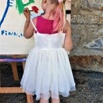 A young girl dressed in a white dress, standing and painting on a wooden canvas outside.