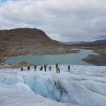 People climbing a blue glacier with a contrasting mountain lake surrounded by bare, snowless mountains in the background.