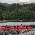 Four people with red life vests in two red canoes on the river, Røssåga, with green forest and mountains in the background.