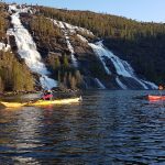 Two people kayaking in front of two big waterfalls in the green mountains behind them.